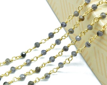 May include: Close-up of multiple strands of gold-colored chain with small, faceted, gray beads. The chains are arranged diagonally across the frame, creating a layered effect. The background is a light-colored, textured surface.