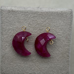 Può includere: Due orecchini a forma di mezzaluna sfaccettati, di colore magenta intenso. Ogni orecchino presenta un anello di filo dorato nella parte superiore per essere appeso. Gli orecchini sono esposti su una superficie grigia.