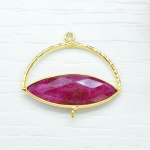 May include: A gold-colored pendant with a large, faceted, oval-shaped, deep red gemstone. The pendant has a loop at the top and a small loop at the bottom. The gemstone is set within a gold frame. The background is white.