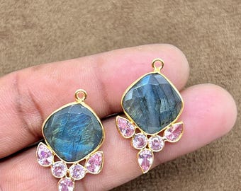 May include: Two gold-toned earrings with a square, faceted, blue-green gemstone center and pink gemstone accents. The earrings have a small loop at the top for hanging. The background is a solid brown color.