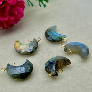 May include: Five faceted labradorite moon-shaped pendants with gold-colored wire loops. The pendants display iridescent colors of blue, green, and yellow. The background is a light beige color, with a pink and white flower in the upper left corner.
