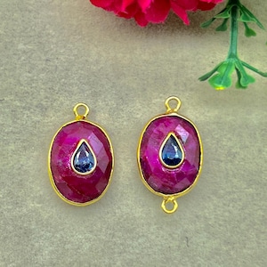 May include: Two oval-shaped pendants featuring faceted magenta-colored stones set in gold-toned metal. Each pendant has a small, dark blue, teardrop-shaped stone embedded in the center. One pendant has a single top loop, while the other has two loops.