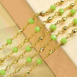May include: Four strands of gold chain with light green glass beads. The chains are layered on top of each other and are lying on a beige surface.