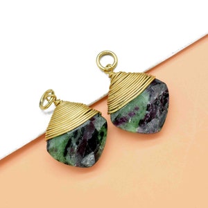 May include: Two green and purple gemstone pendants wrapped in gold wire. The pendants are on a white and peach background.