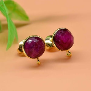 May include: A pair of gold earrings with a deep pink gemstone. The earrings have a small loop on the bottom.