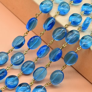 May include: Strands of blue oval-shaped beads with a translucent appearance, connected by delicate gold-colored chains. The beads vary slightly in size and shape, creating a textured look. The background is a soft peach color.