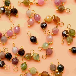 Puede incluir: Un surtido de cuentas de piedras preciosas facetadas en varios colores, incluyendo rosa, verde, marrón y negro, están ensartadas con alambre dorado. Las cuentas están dispuestas sobre una superficie color melocotón, mostrando sus diversos tonos y texturas.