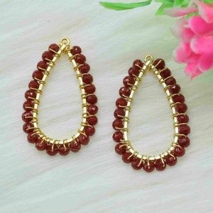 May include: A pair of gold-toned earring blanks with a teardrop shape, adorned with maroon-colored beads.