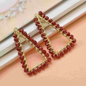 May include: A pair of gold-toned earrings with a geometric triangle shape. The earrings are made with small, red beads.