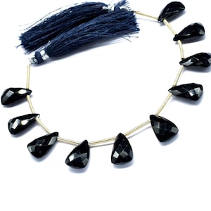 May include: A strand of black gemstone beads with a faceted teardrop shape. The beads are strung on a gold wire and have a tassel at the end.