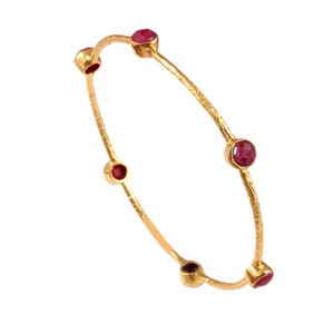 Könnte beinhalten: Ein goldfarbenes Armreif-Armband mit strukturierter Oberfläche. Das Armband hat fünf runde, rote Edelsteine, die gleichmäßig über das Band verteilt sind. Die Edelsteine sind in einer Krappenfassung gefasst.