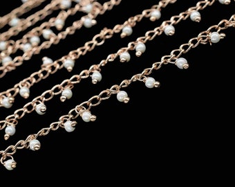 Op de afbeelding: Een close-up van een gouden ketting met kleine witte kralen die eraan hangen. De ketting ligt op een zwarte achtergrond.