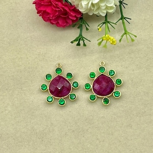 May include: Two gold pendants, each with a central, faceted, deep red gemstone encircled by eight green gemstones. The pendants are set against a neutral backdrop, with red, white, and yellow flowers in the background.