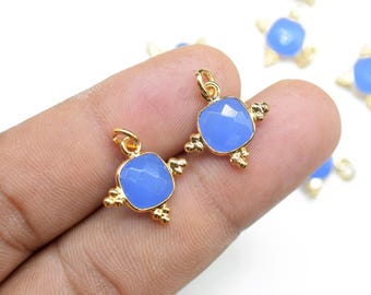 Puede incluir: Dos colgantes dorados con piedras preciosas cuadradas de color azul claro. Cada colgante tiene un pequeño lazo en la parte superior y detalles dorados decorativos. Varios colgantes similares son visibles en el fondo.