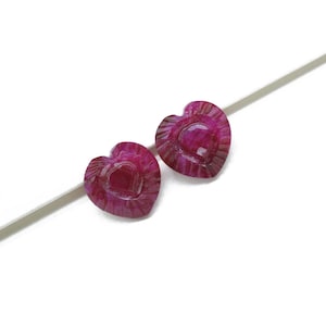 May include: Two heart-shaped, deep magenta gemstones with carved details. The gemstones are set against a white background. The heart-shaped stones have a textured, ridged border and a smooth, central heart shape.