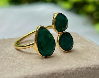 Puede incluir: Un anillo de oro con tres piedras preciosas de color verde esmeralda. La piedra más grande tiene forma de pera, mientras que las otras dos son redondas. La banda del anillo es fina y dorada, y las piedras están engastadas en biseles dorados.