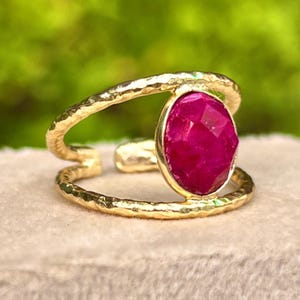 May include: A gold-colored ring with a large, oval-shaped, faceted, magenta-colored gemstone. The ring has a textured band that wraps around the finger. The gemstone is set in a gold bezel.