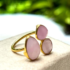 Może przedstawiać: Złoty pierścionek z trzema jasno różowymi kamieniami szlachetnymi. Kamienie są osadzone w złocie i mają fasetowany wygląd. Pierścionek jest prezentowany na neutralnej powierzchni z rozmytym zielonym tłem.