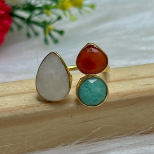 Può includere: Un anello dorato con tre incastonature di pietre preziose: una goccia bianca, una goccia rossa e una pietra turchese rotonda. L'anello è esposto su una superficie di legno.