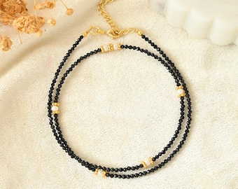 Puede incluir: Un collar de cuentas negras de doble hebra con detalles dorados y pequeñas perlas blancas. El collar tiene una cadena fina y se muestra sobre una superficie color crema. La joya está diseñada para un look sofisticado.