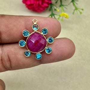 May include: A gold pendant with a central, faceted, magenta gemstone, encircled by eight teal-coloured gemstones. The pendant has a loop at the top for a chain. The gemstones are set in a gold setting.