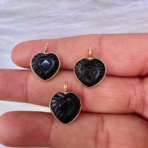 May include: Three black heart-shaped pendants with gold-coloured bails. Each pendant features a carved heart design within a gold-coloured frame. The pendants are displayed against a soft, white background.