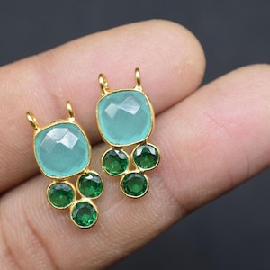 May include: Two gold-toned earrings with a square, faceted, light blue gemstone at the top and three smaller, round, green gemstones below. The earrings are held between fingers against a dark background.