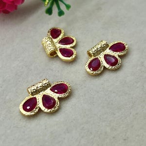 Peut inclure: Trois composants de bijoux dorés en forme de papillon avec des surfaces texturées. Chaque pièce présente trois pierres précieuses rouges profondes en forme de goutte d'eau. Les composants sont disposés sur une surface gris clair, avec des accents floraux en arrière-plan.