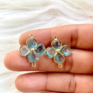 Puede incluir: Dos componentes de joyería dorados en forma de flor de cuatro pétalos. Cada pétalo es una piedra preciosa facetada de color azul claro. Los componentes tienen pequeños bucles para la fijación y un centro dorado.