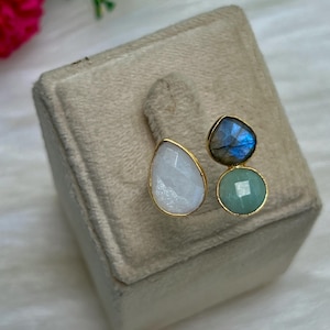 Könnte beinhalten: Drei Ohrringe mit Edelsteinen und goldfarbenen Fassungen werden auf einer beigefarbenen Samtoberfläche präsentiert. Die Ohrringe haben einen tropfenförmigen weißen Stein, einen blau-grauen Stein und einen runden, hellgrünen Stein. Die Ohrringe sind vor einem weichen, weißen Hintergrund platziert.
