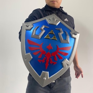 May include: A blue and silver shield with a red emblem and gold accents. The shield has a stylized design with a central emblem and a silver border. The shield is held by a person.