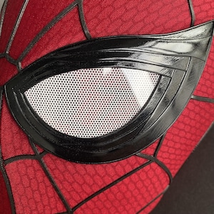 May include: Close-up of a red and black Spider-Man mask. The mask features a black, glossy eye frame with a white, perforated lens. The red fabric has a webbed pattern, with black lines outlining the design. The mask is a superhero costume accessory.