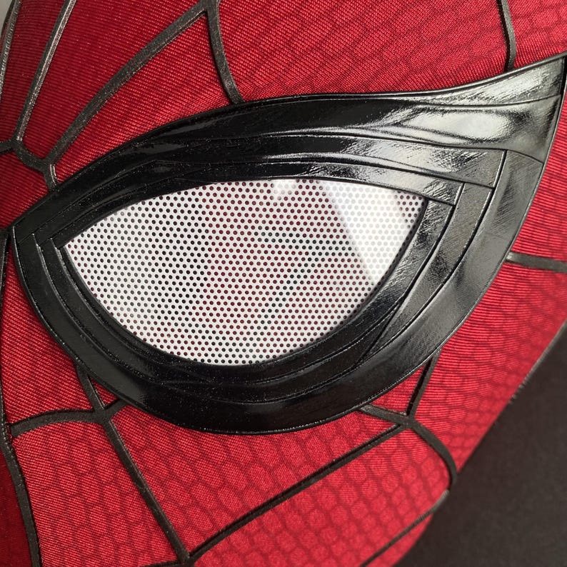 May include: Close-up of a red and black Spider-Man mask. The mask features a black, glossy eye frame with a white, perforated lens. The red fabric has a webbed pattern, with black lines outlining the design. The mask is a superhero costume accessory.