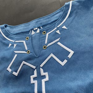 Pu&ograve; includere: Primo piano di una camicia blu con disegni geometrici ricamati in bianco. La camicia ha uno scollo tondo con finiture bianche e una piccola apertura a forma di V. Sono visibili occhielli color oro vicino allo scollo. La camicia sembra essere realizzata in un tessuto morbido e resistente.