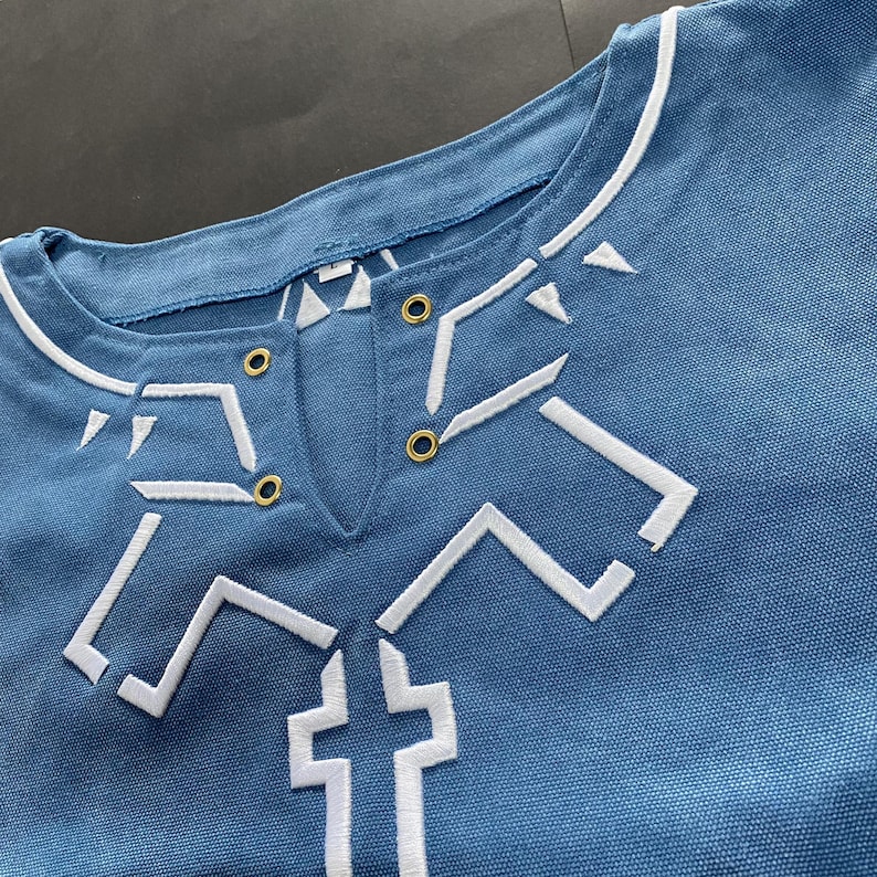 Pu&ograve; includere: Primo piano di una camicia blu con disegni geometrici ricamati in bianco. La camicia ha uno scollo tondo con finiture bianche e una piccola apertura a forma di V. Sono visibili occhielli color oro vicino allo scollo. La camicia sembra essere realizzata in un tessuto morbido e resistente.