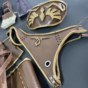 Pu&ograve; includere: Pezzi di armatura cosplay marroni e beige, tra cui un'armatura pettorale e una maschera per il viso. L'armatura ha un design dettagliato con rivetti e forme geometriche. Altri accessori includono una piccola borsa e guanti.