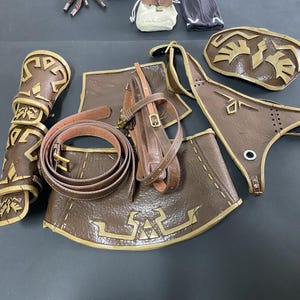 Pu&ograve; includere: Una collezione di pezzi di armatura cosplay marroni e dorati, tra cui protezioni per le braccia, una cintura e armature per petto e gambe. I pezzi presentano intricati disegni geometrici.