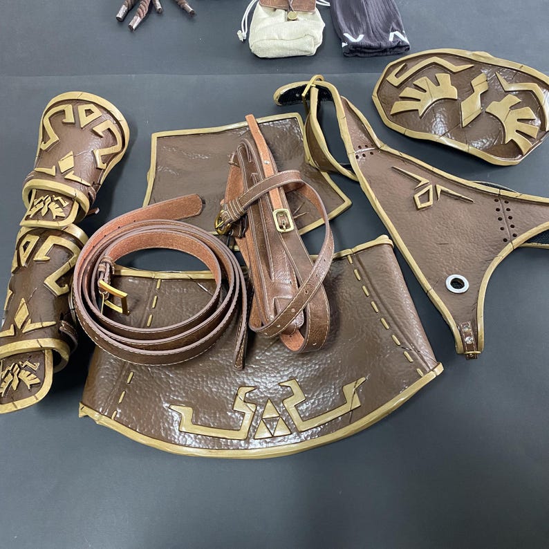Pu&ograve; includere: Una collezione di pezzi di armatura cosplay marroni e dorati, tra cui protezioni per le braccia, una cintura e armature per petto e gambe. I pezzi presentano intricati disegni geometrici.