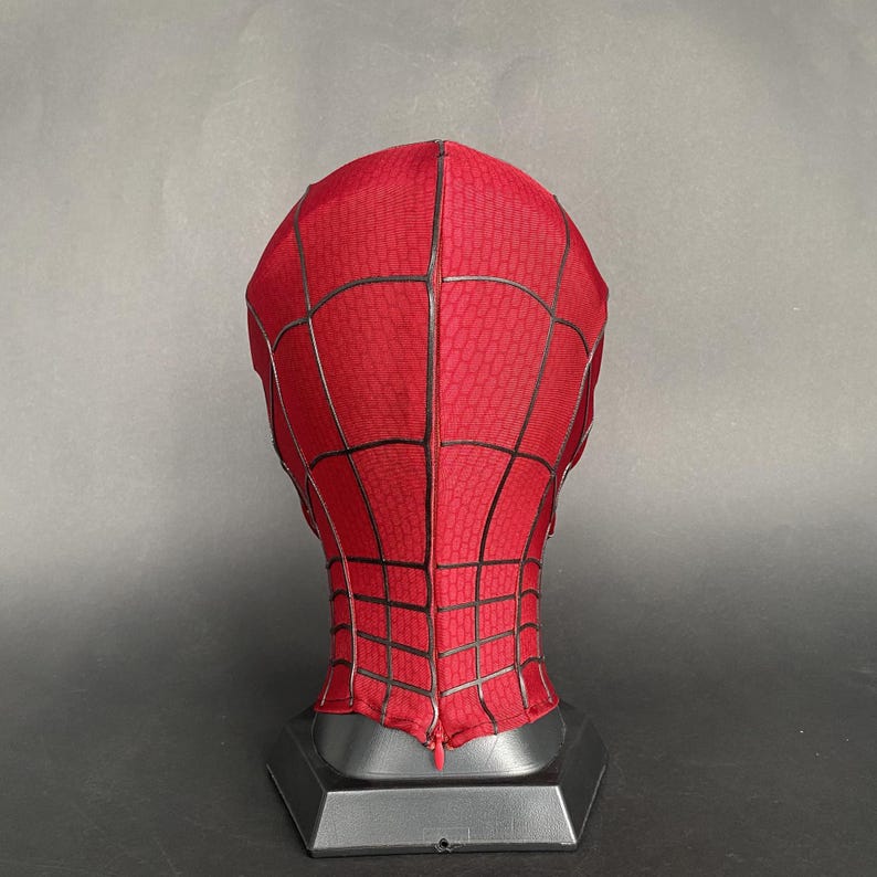 May include: A red Spider-Man mask with a black web pattern. The mask is displayed on a black stand against a gray background. The mask has a textured surface.