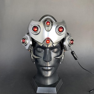 May include: A silver and black helmet with red lights, worn on a mannequin head. The helmet has a futuristic design and is likely a cosplay prop.