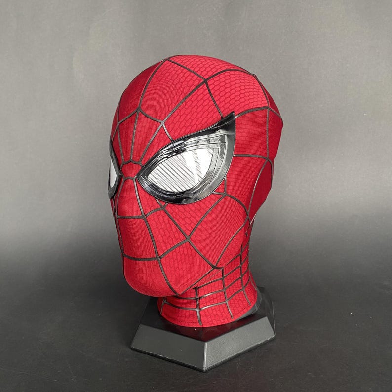 May include: A red and black Spider-Man mask with silver-framed eye lenses. The mask features a web pattern and is displayed on a black stand. The mask is made of a textured material.