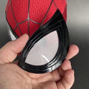 May include: A black, glossy eye lens from a Spider-Man mask, held in a hand. The lens has a white, mesh-like interior and is set against a red and black Spider-Man mask.