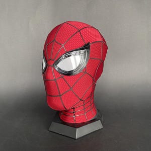 May include: A red and black Spider-Man mask with silver eye lenses. The mask is displayed on a black stand against a gray background. The mask has a textured pattern and black web lines.