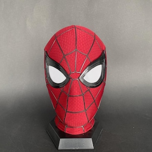 May include: A red and black Spider-Man mask with white eye lenses. The mask has a textured pattern and is displayed on a black stand. The mask is a replica of the superhero's mask.