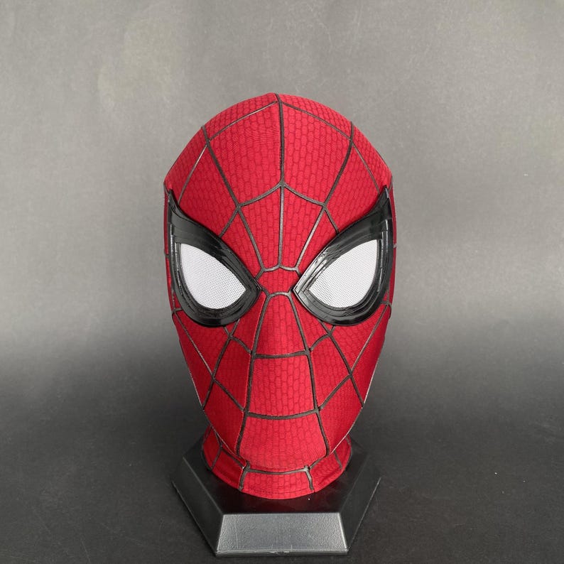 May include: A red and black Spider-Man mask with white eye lenses. The mask has a textured pattern and is displayed on a black stand. The mask is a replica of the superhero's mask.