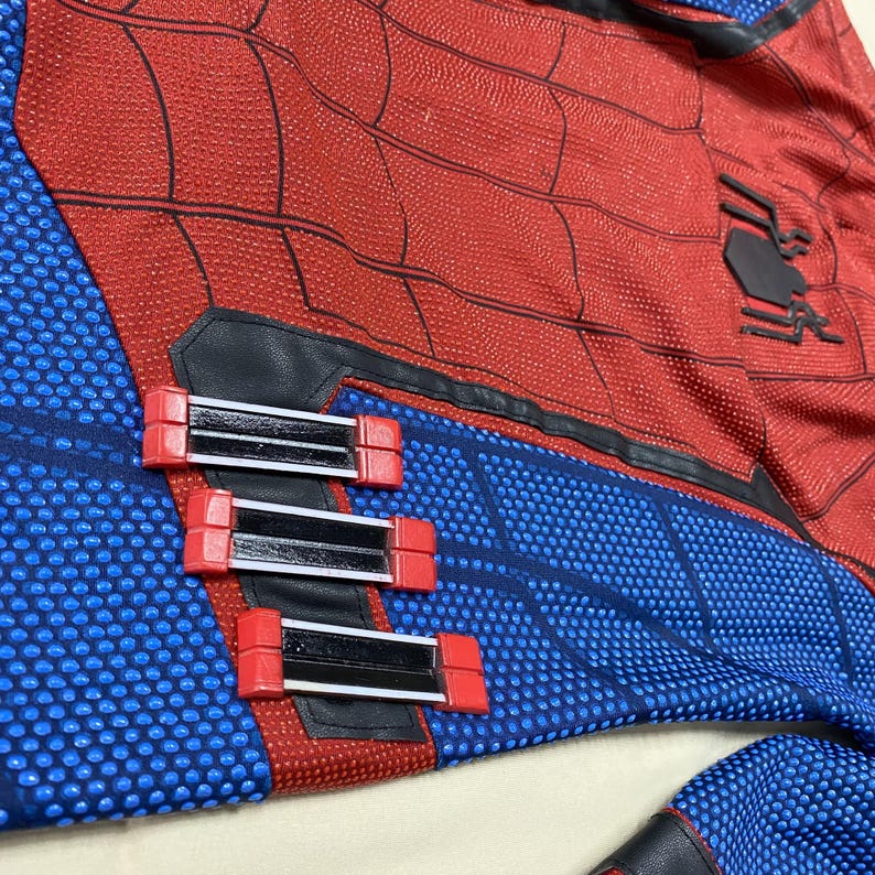 May include: Close-up of a red and blue Spider-Man costume. The costume features a red textured pattern with black lines, blue sections with a dot pattern, and black accents. Three red and black wrist devices are visible.
