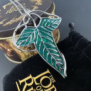 May include: A silver and green enamel pendant necklace featuring a leaf design. The necklace is displayed on a black velvet pouch with the words "The Lord of the Rings" printed on it.