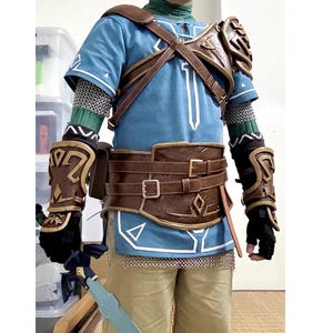 Pu&ograve; includere: Un costume cosplay ispirato a un personaggio di un videogioco. L'outfit include una tunica blu con dettagli bianchi, un'armatura in pelle marrone, una cintura e dei guanti. Il costume presenta anche una cotta di maglia e una spada.