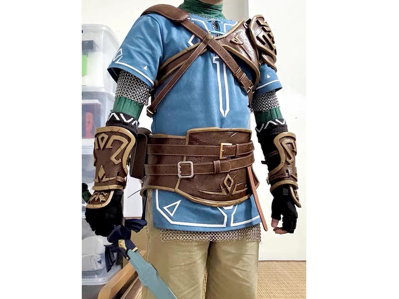 Pu&ograve; includere: Un costume cosplay ispirato a un personaggio di un videogioco. L'outfit include una tunica blu con dettagli bianchi, un'armatura in pelle marrone, una cintura e dei guanti. Il costume presenta anche una cotta di maglia e una spada.
