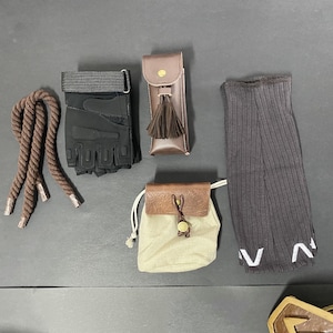 Pu&ograve; includere: Un assortimento di accessori tra cui una corda marrone, guanti senza dita neri, un astuccio in pelle marrone con nappina, una piccola borsa con coulisse beige e marrone e scaldamuscoli a coste grigio scuro con scritte bianche.